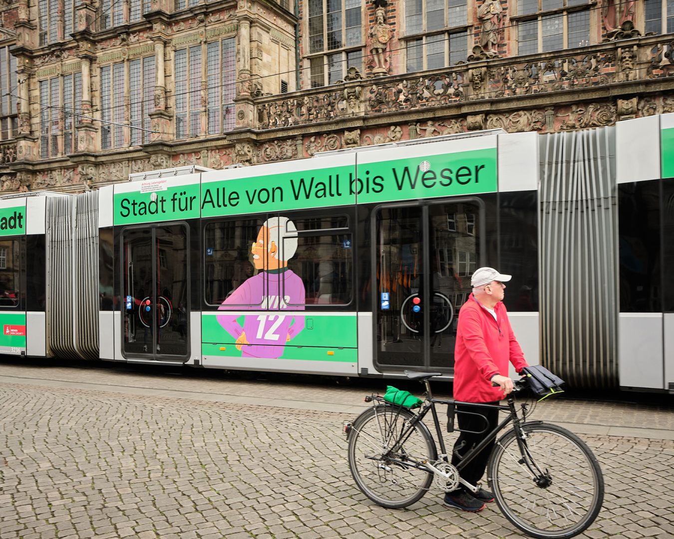Eine Person mit Fahrrad schaut auf eine vorbeifahrende Straßenbahn mit Kampagne „binnenstadt – Stadt für alle“