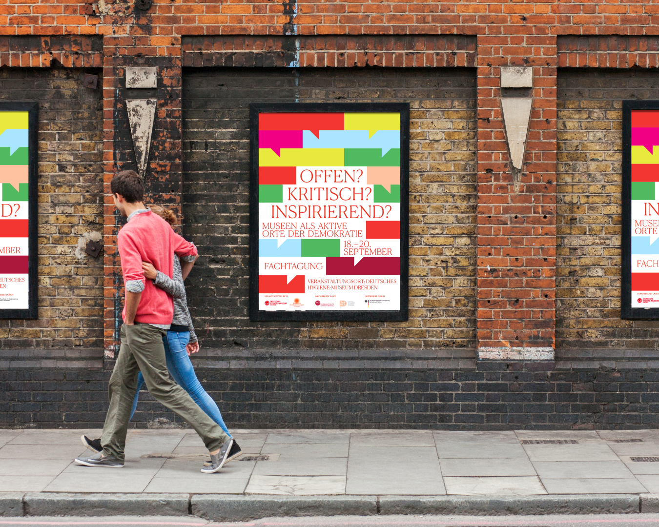 Straßenszene mit drei nebeneinander hängenden Plakaten an einer Backsteinwand. Auf den Plakaten steht in roter Schrift: „Offen? Kritisch? Inspirierend? Museen als aktive Orte der Demokratie. 18.–20. September. Fachtagung. Veranstaltungsort: Deutsches Hygiene-Museum Dresden.“ Darüber bunte grafische Sprechblasen in Grün, Pink, Gelb, Blau und Rot. Im Vordergrund geht ein Paar Arm in Arm an den Plakaten vorbei.