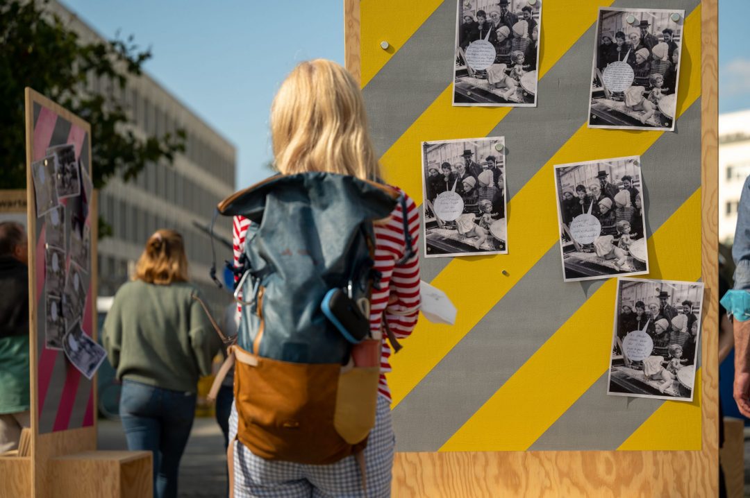 Foto der mobilen Ausstellung „Geschichte unterwegs“ auf dem eine Besucherin historische Fotos mit Zitaten auf farbiger Tafel im Außenraum betrachtet.