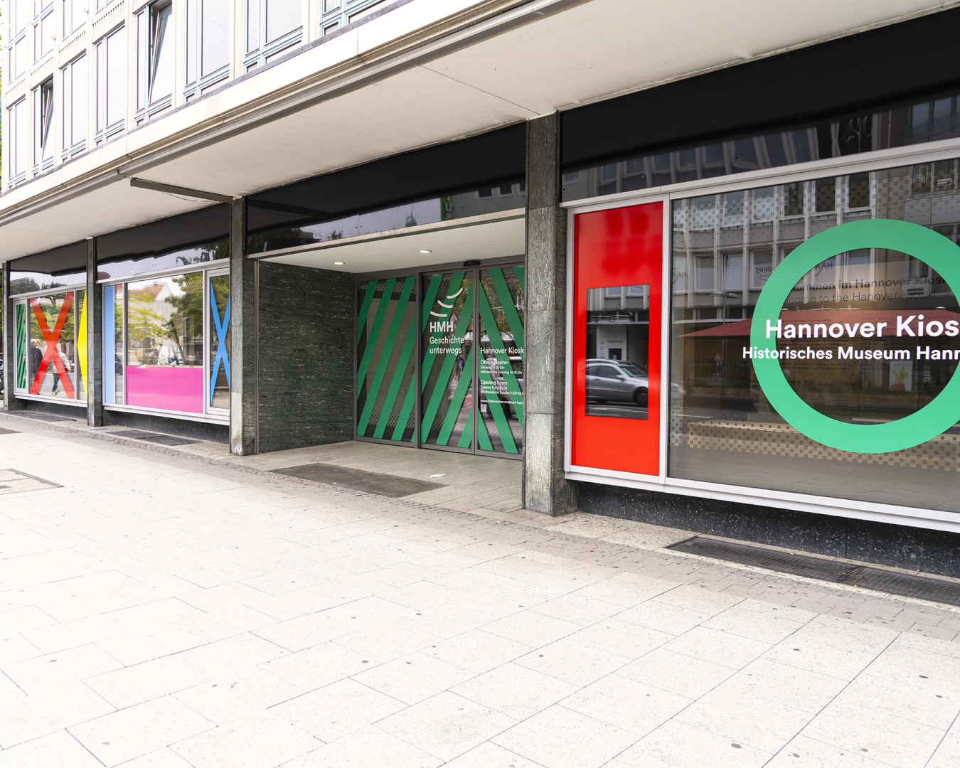 „Hannover Kiosk“ des Historischen Museums Hannover mit farbiger Fensterbeklebung und grünem Logo im öffentlichen Stadtraum.
