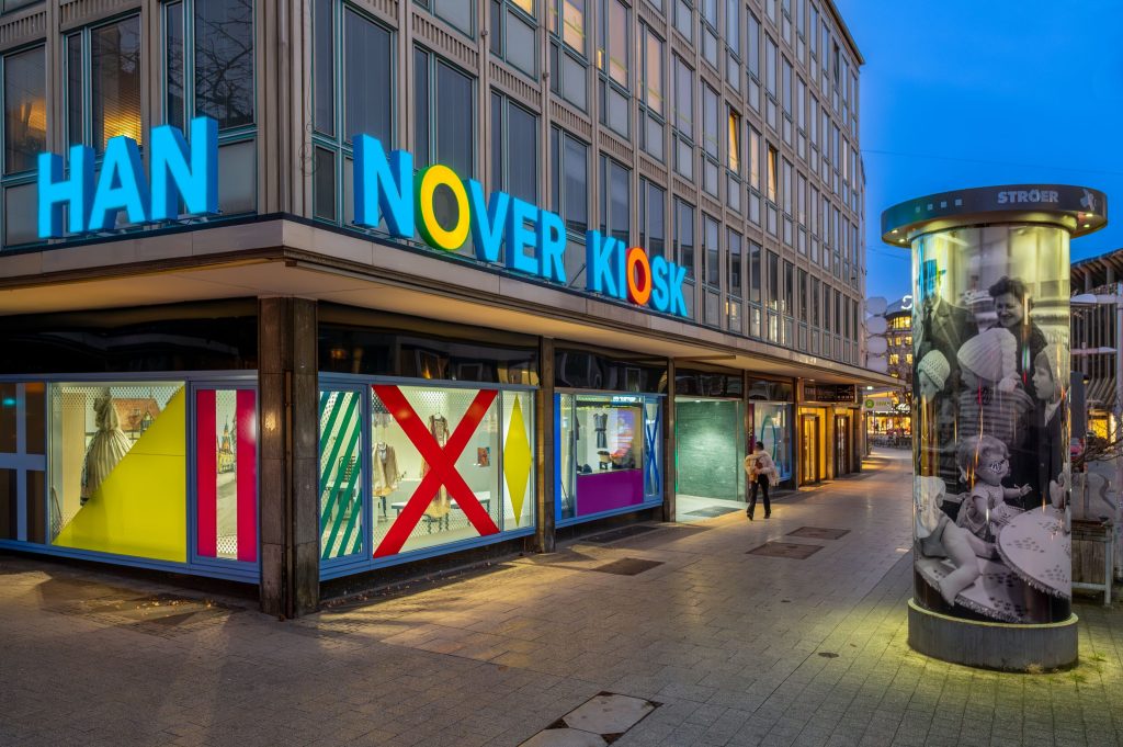 Abendliche Straßenszene in Hannover mit beleuchtetem Eckgebäude, auf dem in großen bunten Buchstaben „HANNOVER KIOSK“ steht. Die Schaufenster sind auffällig gestaltet mit farbigen Flächen, Streifen und geometrischen Formen. Rechts im Bild eine Litfaßsäule mit historischen Schwarz-Weiß-Fotografien. Eine Person geht auf dem Gehweg vorbei, die Straße ist ruhig belebt.