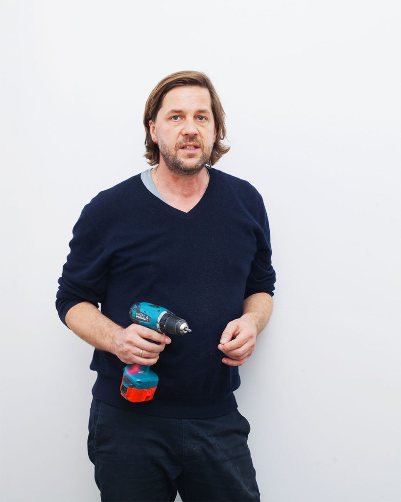 Eine Person mit Akkuschrauber in der linken Hand steht vor weißer Wand