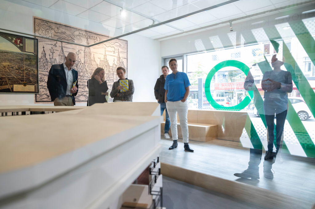 Innenaufnahme des Ausstellungsraums im Hanover Kiosk mit mehreren Personen, die sich um ein großes Stadtmodell versammeln. Im Hintergrund hängen historische Stadtpläne von Hannover an der Wand. Der Raum ist hell und modern gestaltet, durch die mit grafischen Elementen beklebte Fensterscheibe fällt Tageslicht. Auf der Fensterscheibe steht spiegelverkehrt „HANNOVER KIOSK“.
