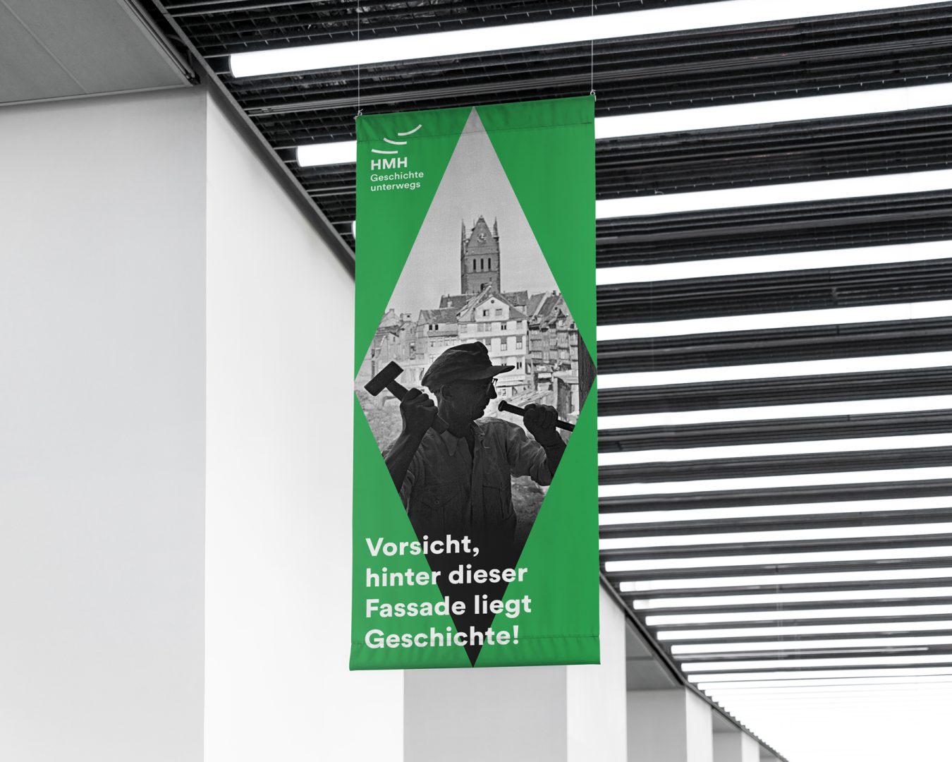 Bannerdesign mit der Aufschrift "Hinter dieser Fassade liegt Geschichte". Das Banner ist grün mit einer Raute in der MItte in der ein schwarz-weiß Foto liegt auf dem ein Mann mit einem Hammer eine Fassade bearbeitet.
