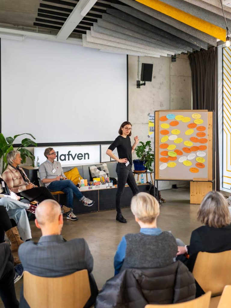 Tagungsszene in einem modernen Raum: Eine Frau steht vorn und spricht vor einer Gruppe sitzender Personen. Neben ihr ein Flipchart mit vielen farbigen Moderationskarten. Im Hintergrund eine leere Projektionsfläche, Sitzbank mit Materialien und das Logo „hafven“. Die Atmosphäre wirkt konzentriert und partizipativ.