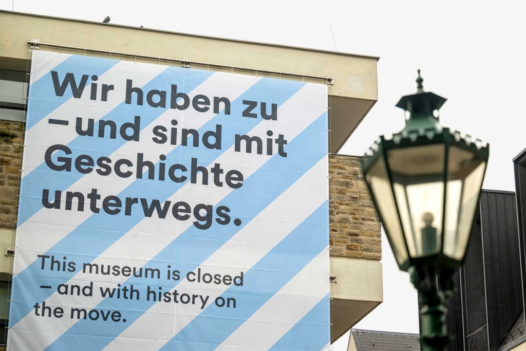 Großes Banner an der Fassade eines Museums mit diagonalem Streifenmuster in Hellblau und Weiß. Der Text darauf informiert zweisprachig über eine Schließung: „Wir haben zu – und sind mit Geschichte unterwegs.“ Darunter auf Englisch: „This museum is closed – and with history on the move.“ Rechts im Vordergrund unscharf eine historische Straßenlaterne.