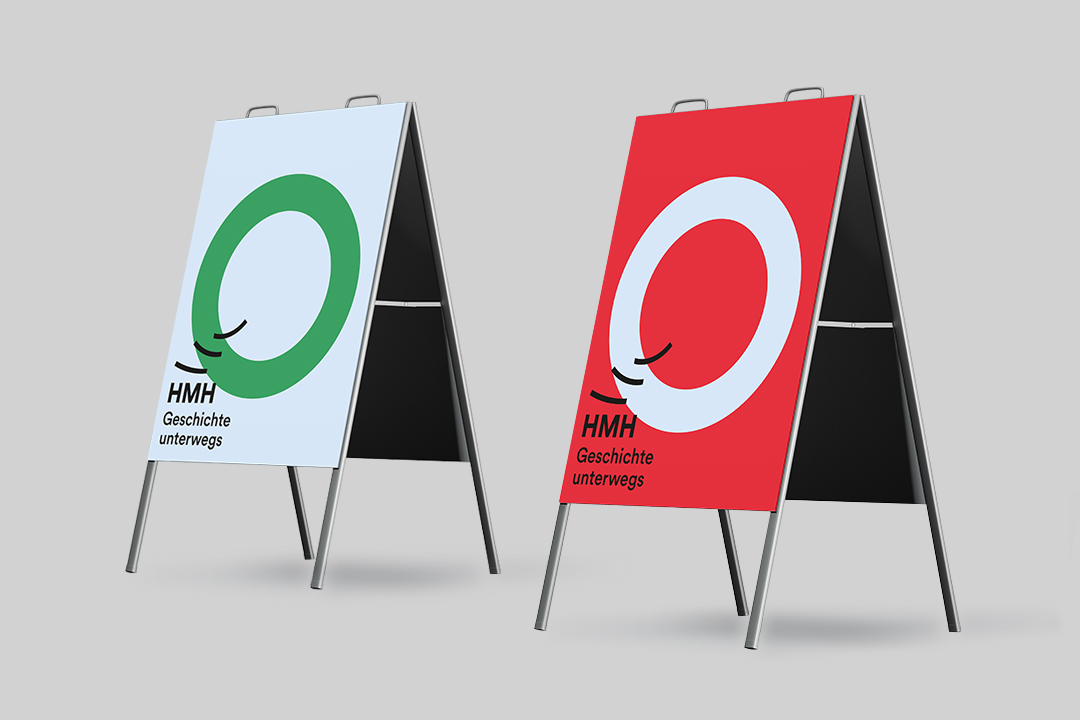 Zwei aufgestellte Kundenstopper-Schilder mit dem Logo „HMH – Geschichte unterwegs“. Links hellblauer Hintergrund mit großem grünem Kreis, rechts roter Hintergrund mit großem weißem Kreis. Unterhalb des Kreises jeweils der schwarze Schriftzug „HMH Geschichte unterwegs“.