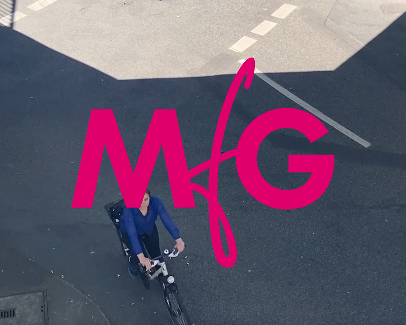 Straßenszene von oben: eine Frau fährt auf einem Fahrrad von oben nach unten durchs Bild. Im Bild ist zentral groß MfG geschrieben.
