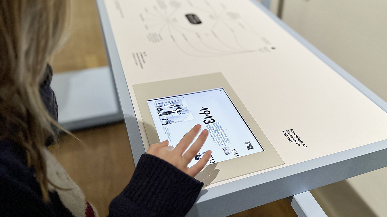Blick über die Schulter eines Kindes, das einen Touchscreen bedient. Darauf eine Zeitleiste mit der Jahreszahl 1913 und Informationstext.