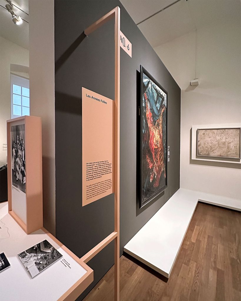 Perspektivisch stark verzerrte Ausstellungsansicht mit koralle-farbendem Dislplay mit Informationstext „Les Années Folles“ und dem Schild „No. 6“