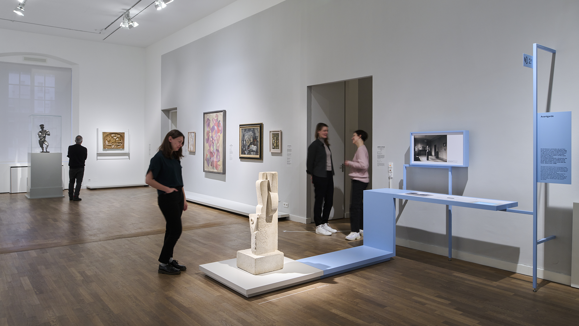 Vier Personen stehen in einem Raum mit Ausstellungsobjekten, betrachten die Kunst und unterhalten sich. Im Vordergrund wird eine Skulptur angeleuchtet.