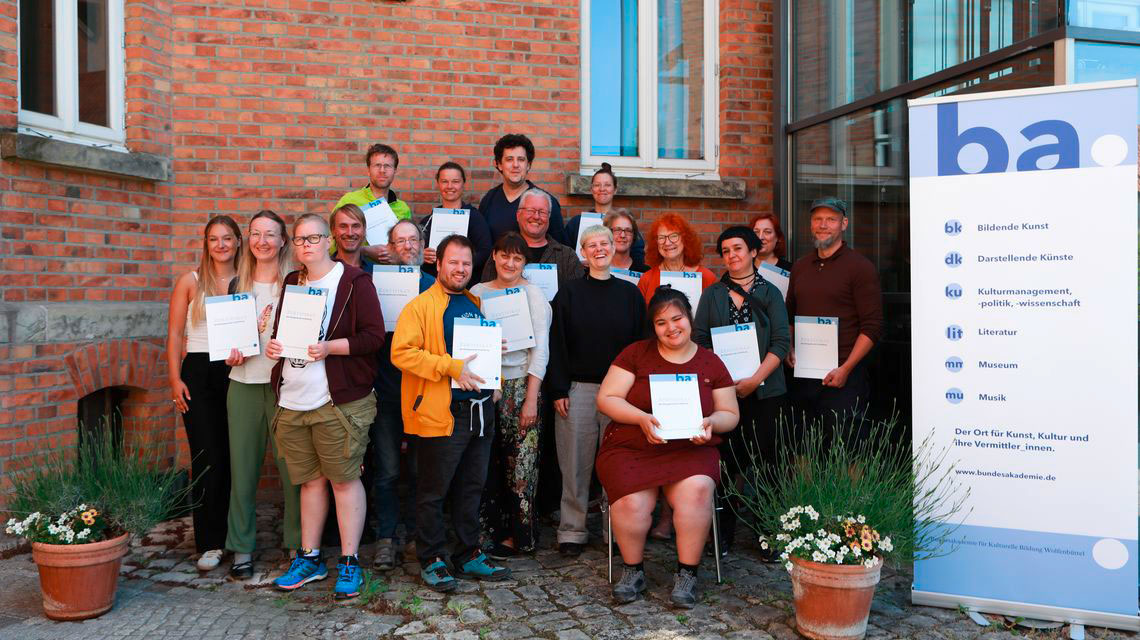Gruppenbild mit Urkunden neben Roll-up der Bundesakademie Wolfenbüttel vor Hauswand mit Backsteinarchitektur und Pflanzen