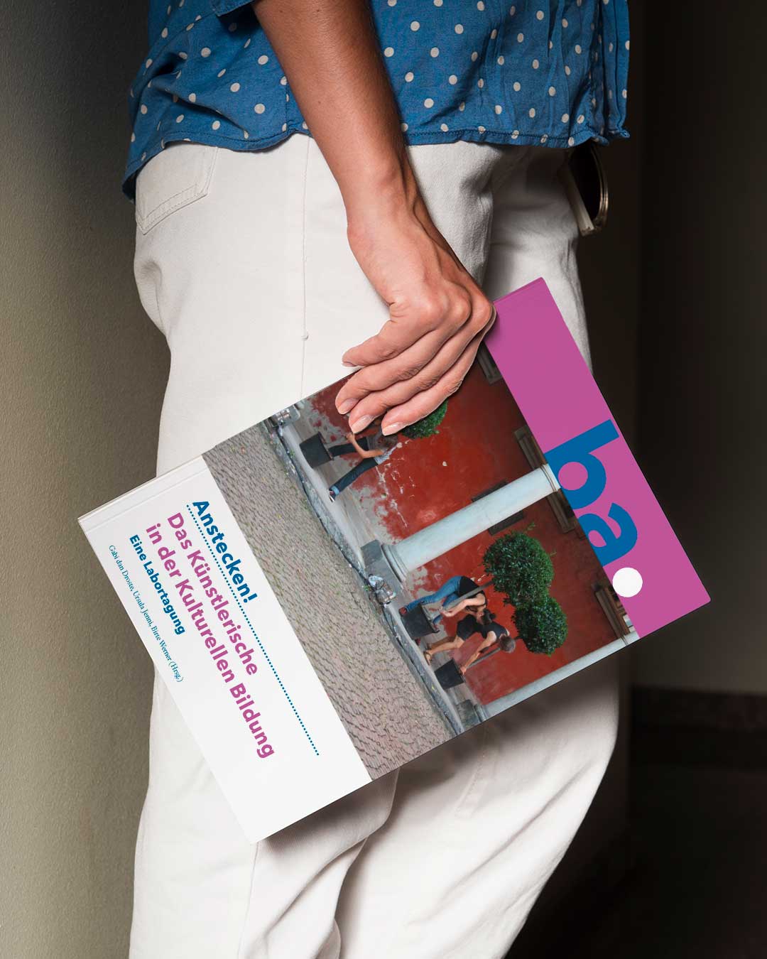 Seitliche Ansicht einer Person. Beinbereich. Person hält eine Publikation im Format DIN A4 mit dem Titel ba