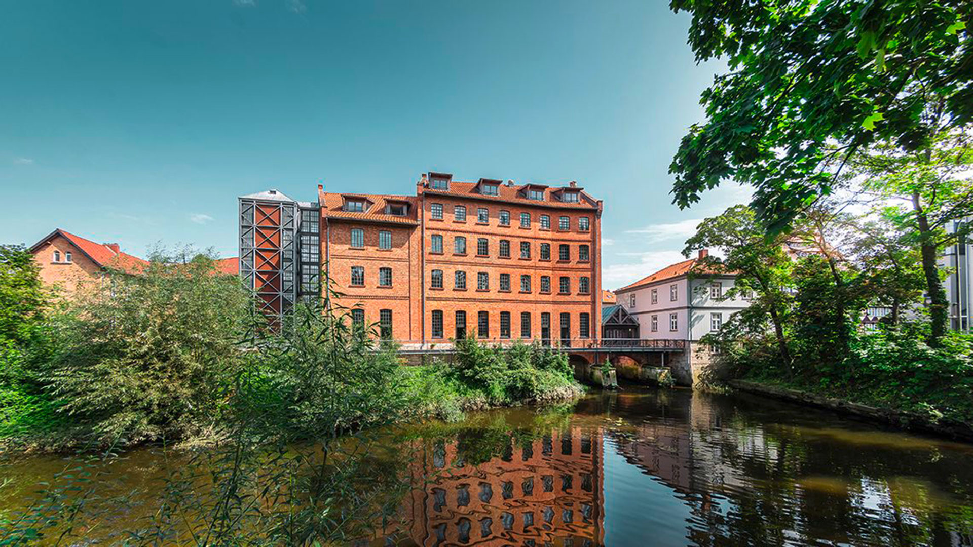 Gästehaus der Bundesakademie Wolfenbüttel. Blick über Wasser. Im Hintergrund die Rückseite des Gästehauses. Roter Backsteinbau. Grüne Umgebung mit Bäumen.