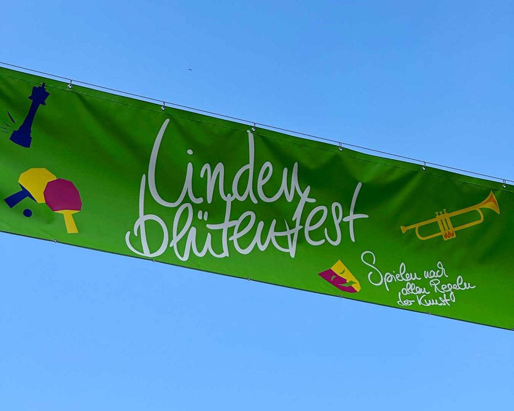 Banner vor blauem Himmel: Lindenblütenfest, weiß auf grün. Piktogramme: Schachfigur Dame, Ping Pong Schläger und Ball, Trompete, Maske. Dazu Schriftzug: Spielen nach allen Regeln der Kunst