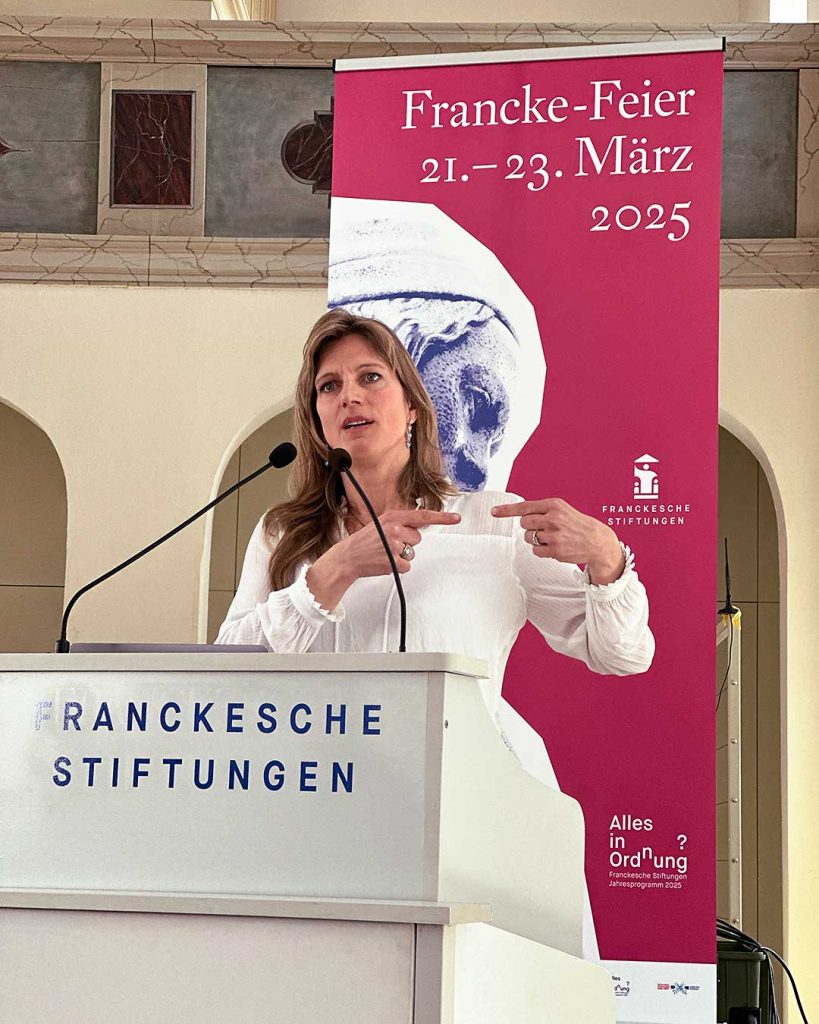 Eine Person auf dem Podium gestikuliert vortragend mit den Zeigefingern gegeneinander. Dahinter Roll-up: Francke Feier