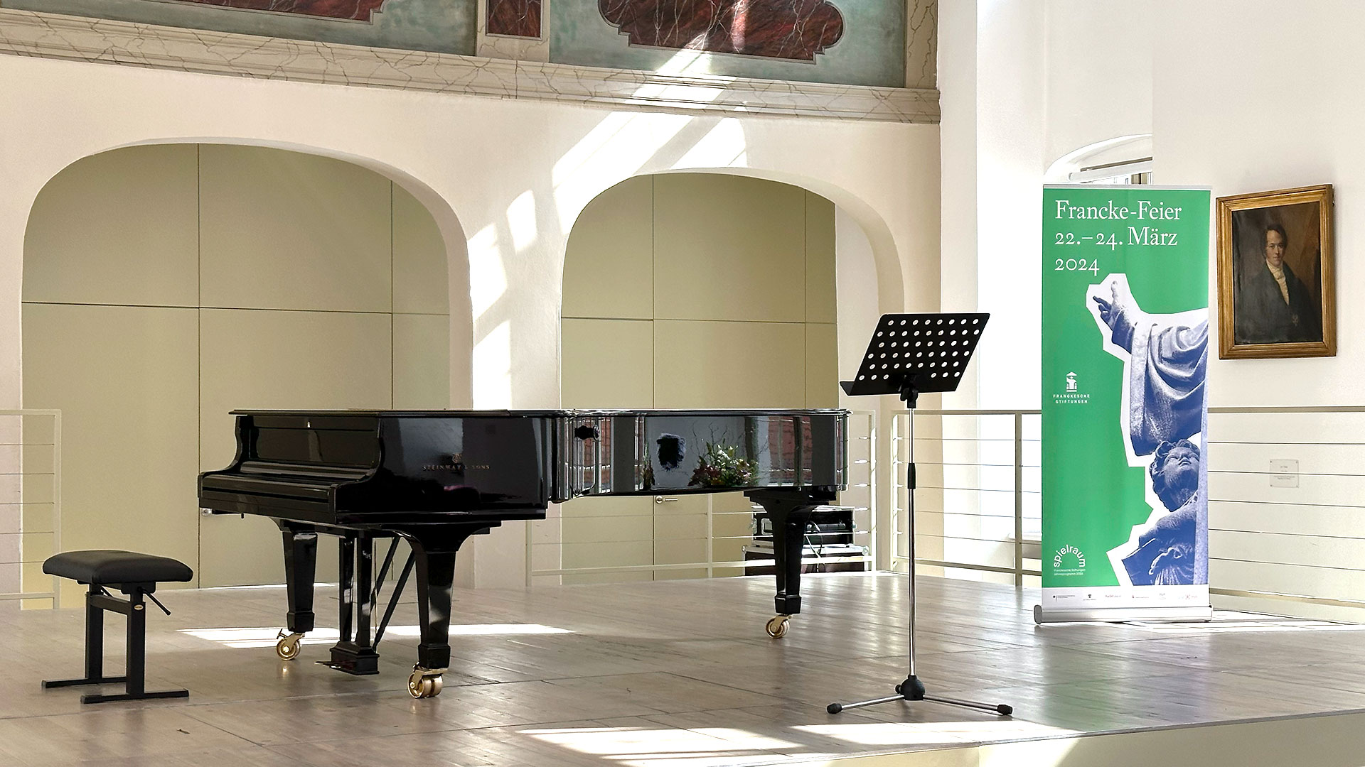 Bühnenansicht Freylinghausen Saal der Franckischen Stiftungen mit Flügel, Notenständer und Roll-up: Francke Feier 2024 in Mintgrün. Darauf Motiv des Francke-Denkmals. Francke Figur weist in den Himmel. Kind schaut auf.