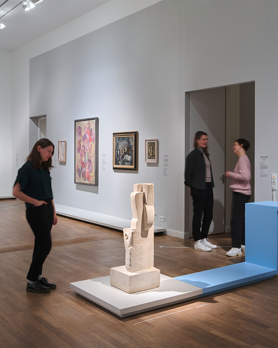 Vier Personen stehen in einem Raum mit Ausstellungsobjekten, betrachten die Kunst und unterhalten sich. Im Vordergrund wird eine Skulptur angeleuchtet.