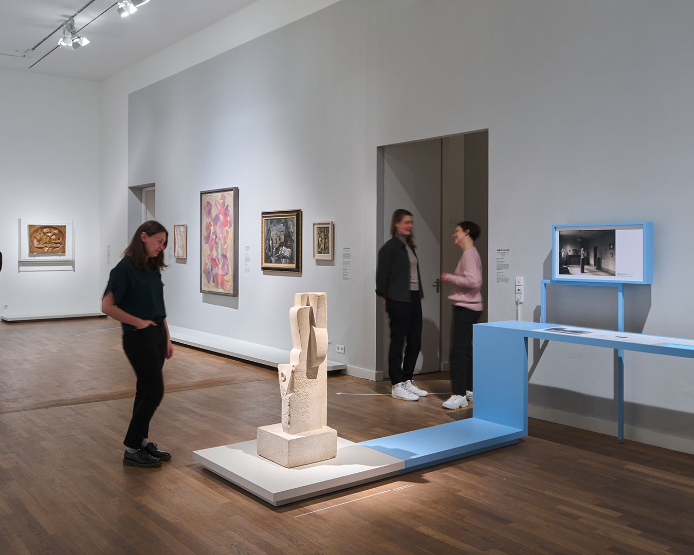 Vier Personen stehen in einem Raum mit Ausstellungsobjekten, betrachten die Kunst und unterhalten sich. Im Vordergrund wird eine Skulptur angeleuchtet.