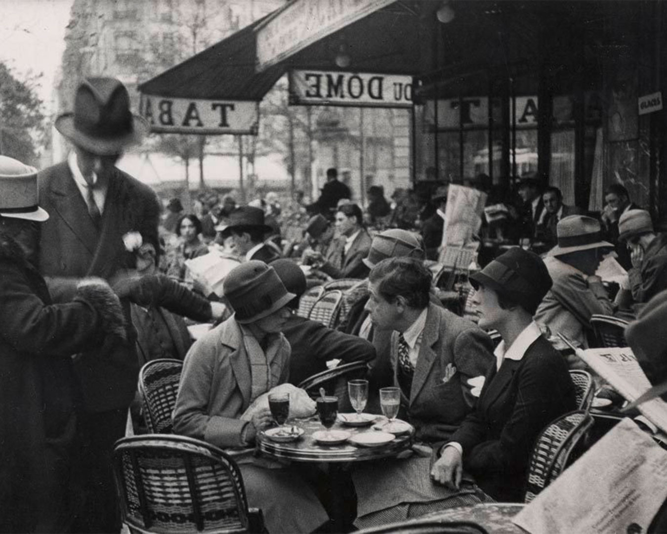 Straßenszene mit Café und Gästen auf historischem Foto. Im Vordergrund unterhalten sich zwei Personen an einem Café-Tisch. Links ein Personenpaar im Gespräch. Im Hintergrund ist der Schriftzug „Café du Dome“ zu erkennen.