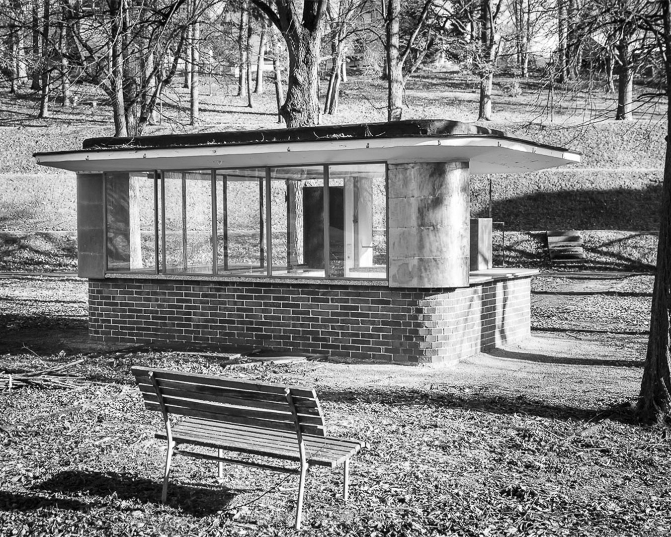 Historisches Foto von einem Kiosk-Gebäude aus Backstein, Glas und Beton. Im Hintergrund bewaldetes Gebiet. Im Vordergrund eine Parkbank.