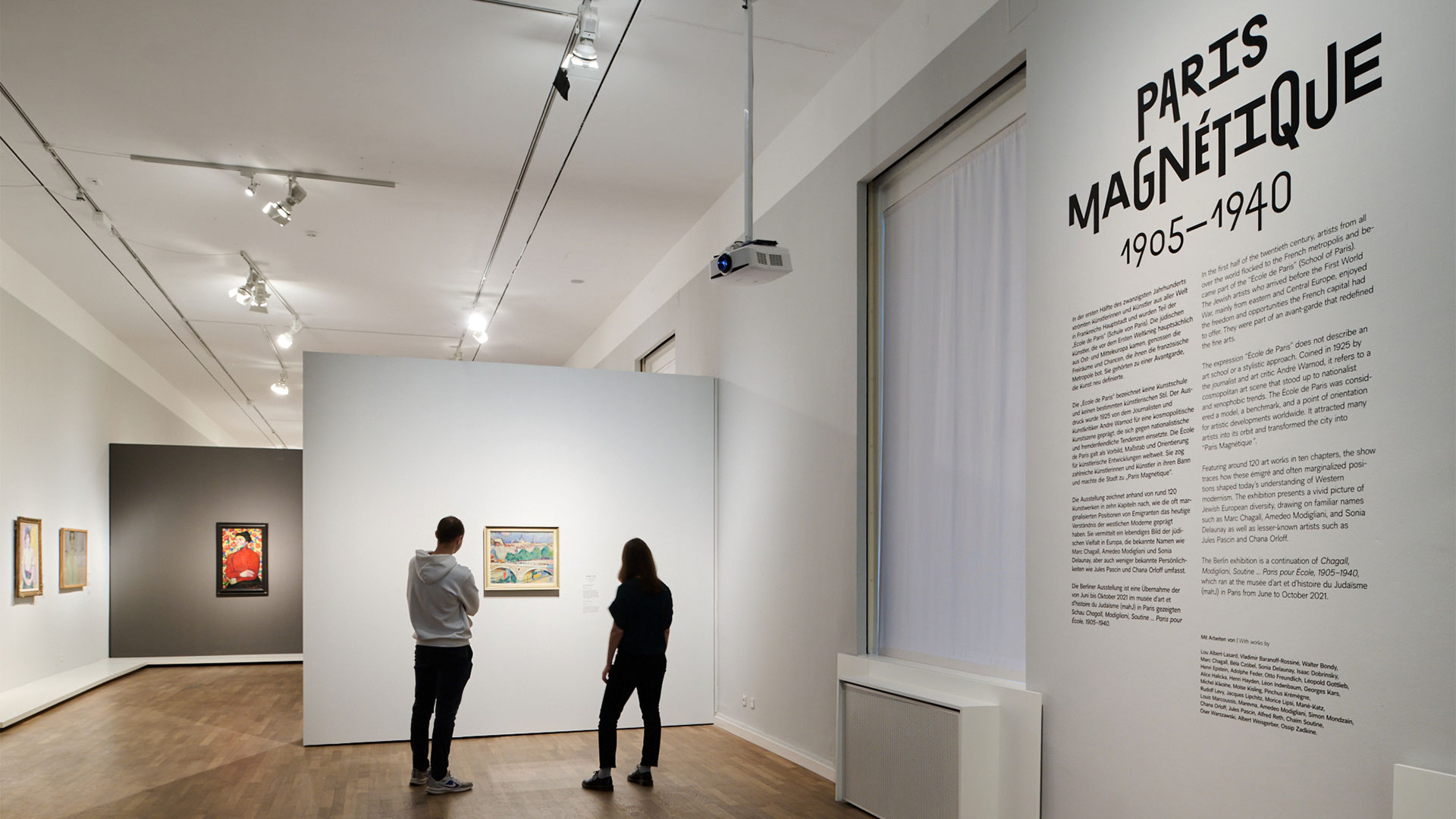 Zwei Personen stehen inmitten eines Ausstellungsraums und betrachten ein Gemälde. Rechts der Ausstellungstext mit der Überschrift „Paris Magnétique 1905-1940“ in Schwarz auf weißer Wand.