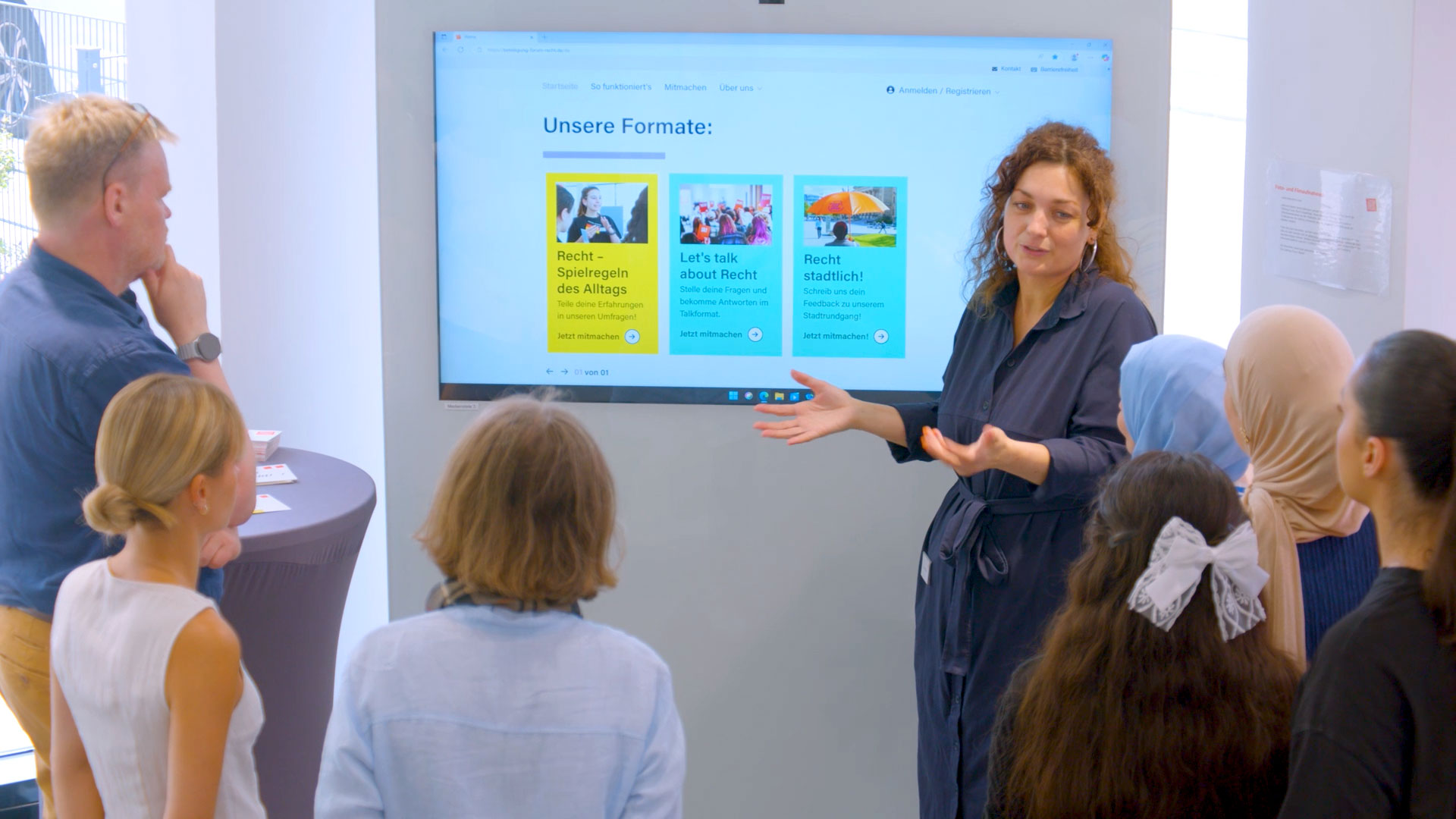 Eine sprechende Person vor einer Präsentation. Rund herum stehen Zuhörende