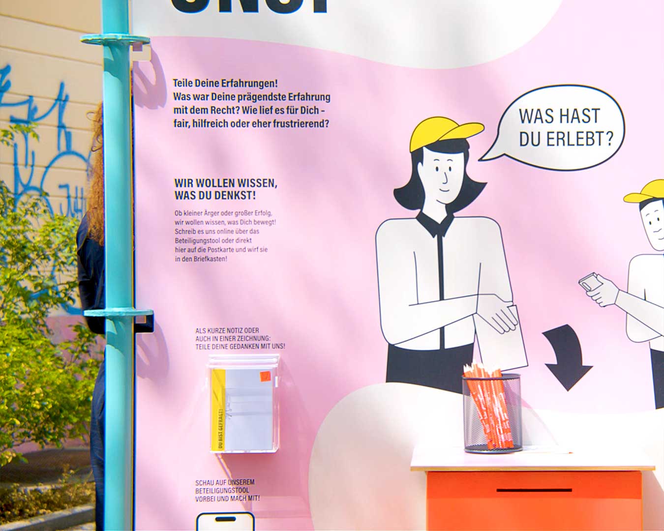 Tafel einer Ausstellung mit Feedback-Aufforderung. Postkarten stehen in einer Box.