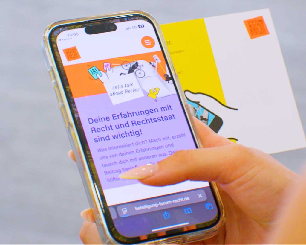 Eine Person hält ein Smartphone, auf der die Kampagnenwebsite der Stiftung Forum Recht angezeigt wird. Dahinter eine Postkarte mit QR-Code.