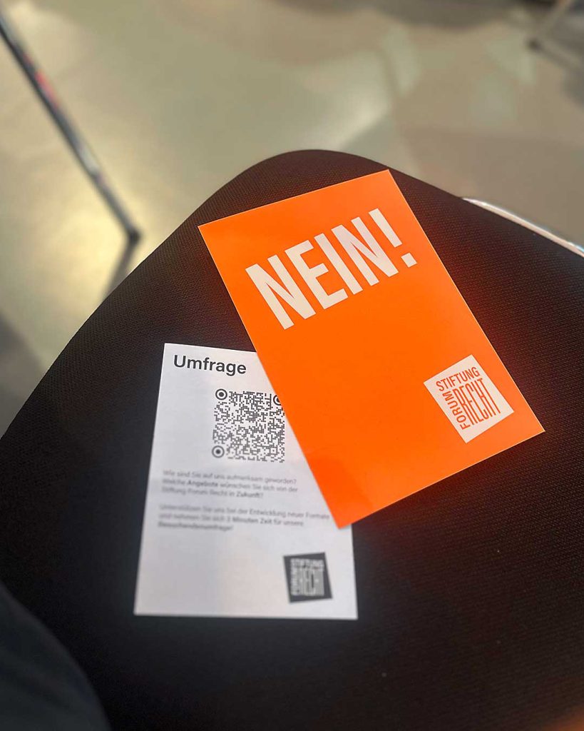 Blick auf eine Sitzfläche eines Stuhls. Schwarzes Polster. Darauf eine Abstimmungskarte, neonorange: „NEIN!“ Logo Stiftung Forum Recht. Daneben Zettel mit QR-Code.