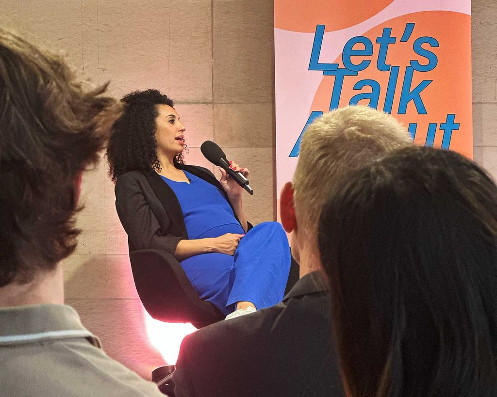 Bühnenansicht durch Publikum. Auf der Bühne eine sprechende Person mit Mikrofon.