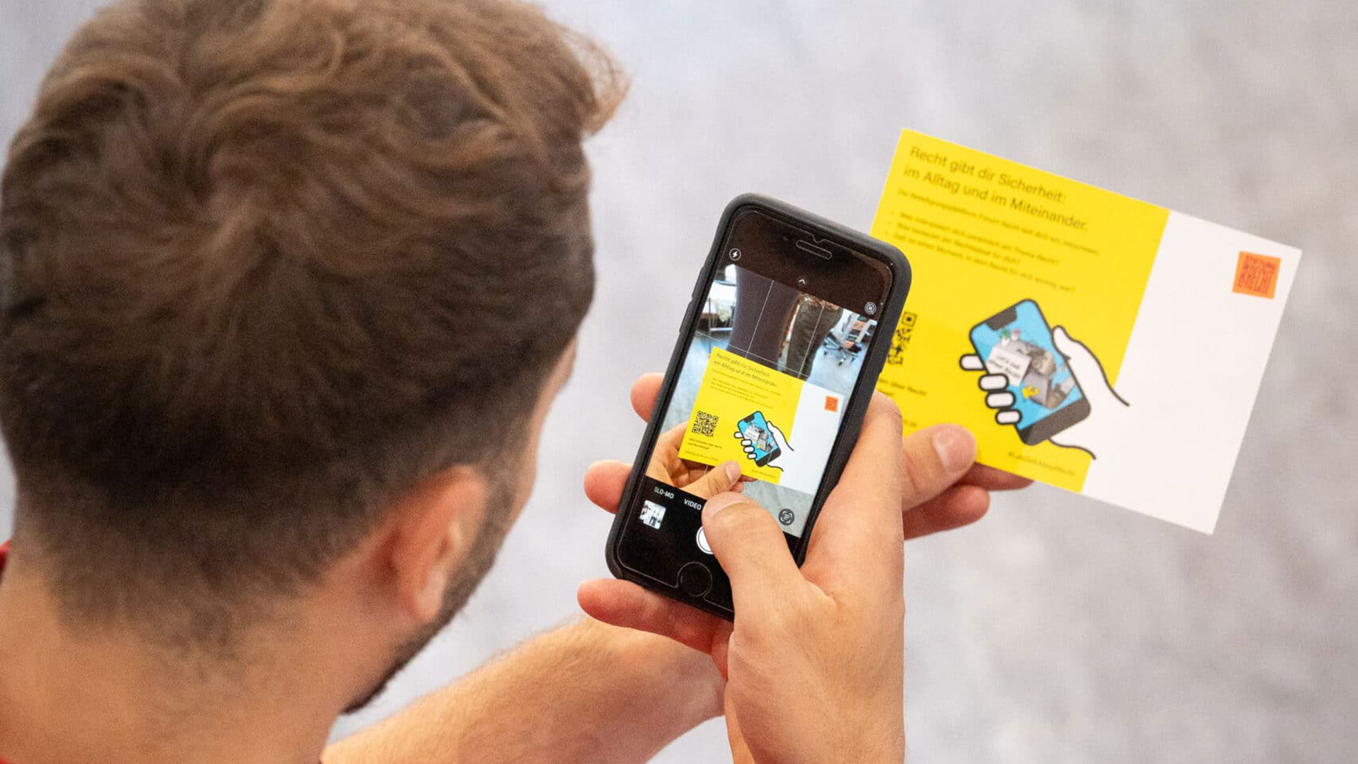Blick über die Schulter einer Person mit Smartphone. Website Beteiligung Formum Recht. Dahinter eine Postkarte mit QR-Code