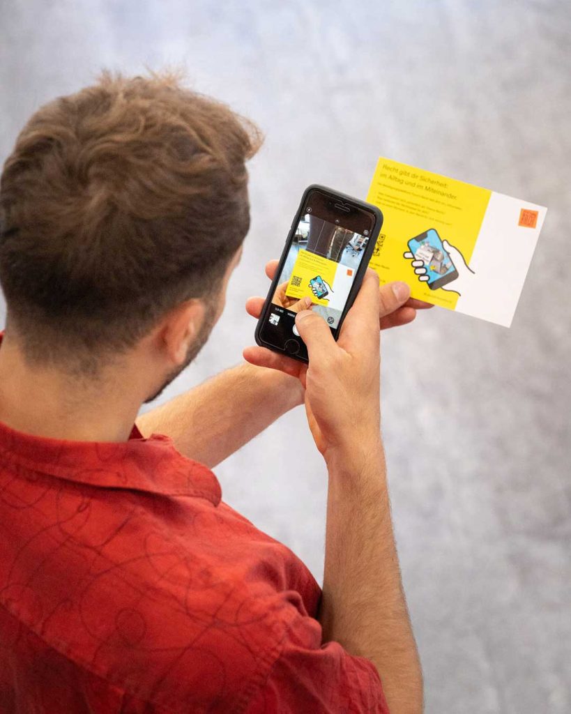 Blick über die Schulter einer Person mit Smartphone. Website Beteiligung Formum Recht. Dahinter eine Postkarte mit QR-Code