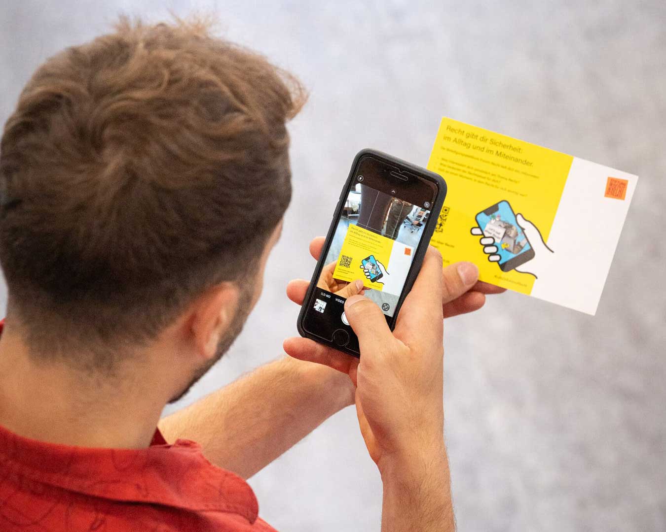 Blick über die Schulter einer Person mit Smartphone. Website Beteiligung Formum Recht. Dahinter eine Postkarte mit QR-Code