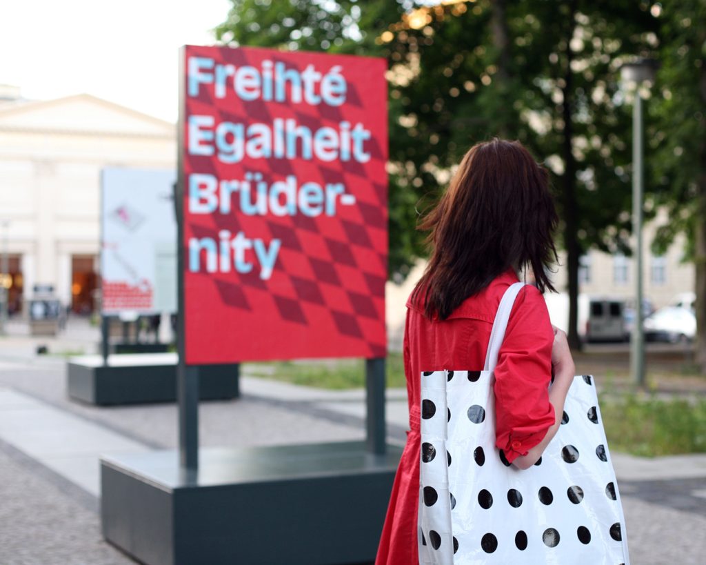 Eine dem Blick abgewandte, braunhaarige Person mit weißer Tasche schaut auf ein Plakat im Hintergrund: „Freihté, Egalheit, Brüdernity“