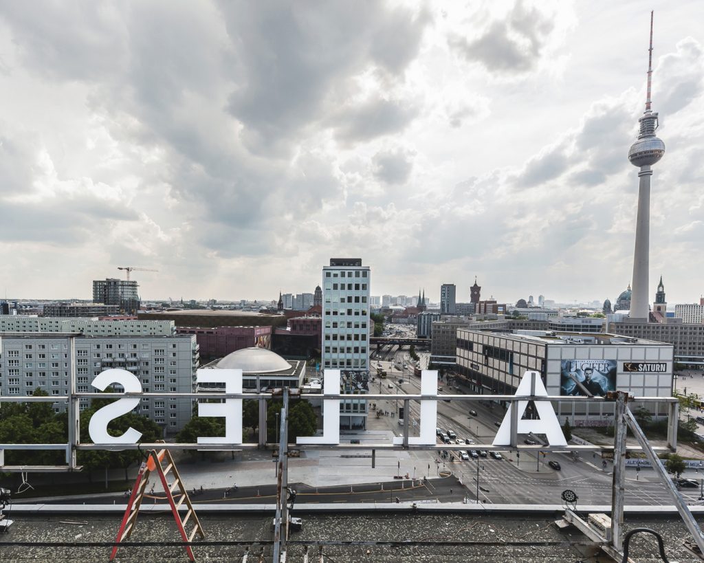 Blick über den Berliner Alexanderplatz vom Dach eines Hochhauses. Von hinten sind weiße Werbebuchstaben „ALLES“ zu erkennen.