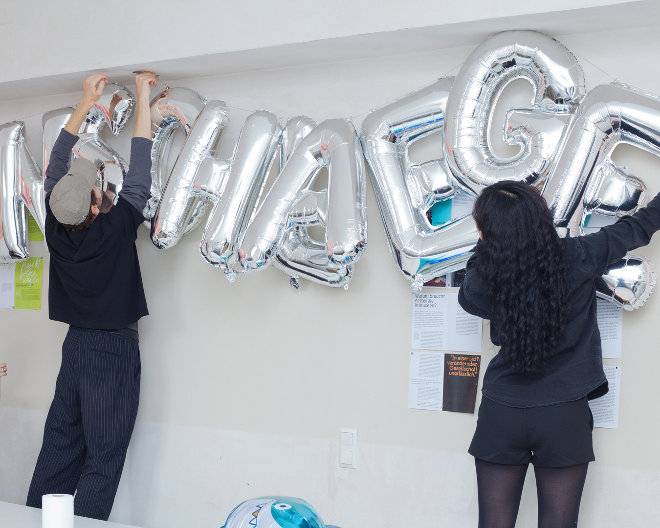 Zwei Personen stehen in Rückenansicht an einer grauen Studiowand und bringen silberne Ballonbuchstaben an: ANSCHLAEGE