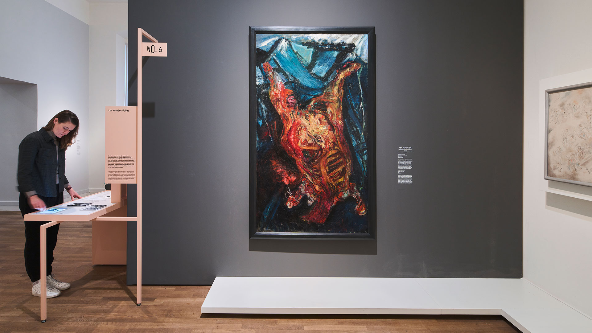 Frontalansicht eines Kunstwerks auf dunkelgrauer Wand. Darauf ein Ölgemälde, dass eine orange-rote Figur vor blauem, zerklüfteten Hintergrund zeigt. Links steht eine Person an einem koralle-farbenen Ausstellungsdisplay und erkundet die Informationen.