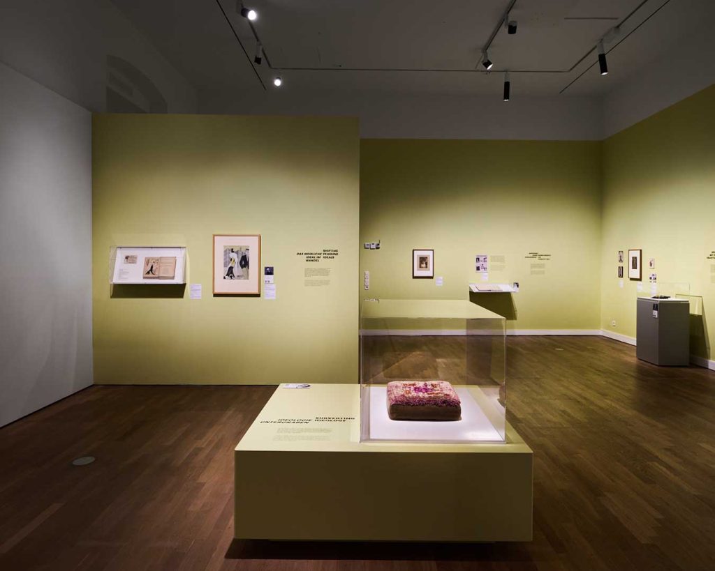 Raumansicht Ausstellung Widerstände. Werke auf gelben Wänden. In der Mitte frontal ein Podest mit Vitrine.