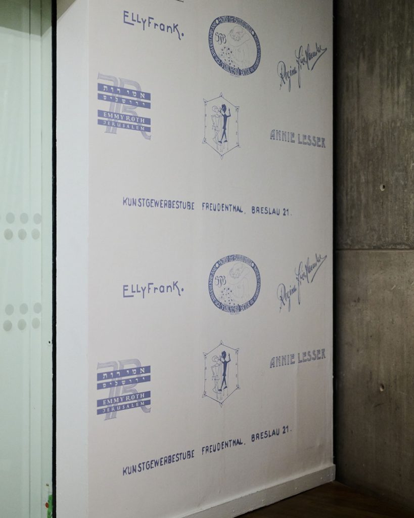 Raumansicht Ausstellung Widerstände. Wandtapete mit historischen Signets der Designerinnen. Teilweise in hebräischer Schrift