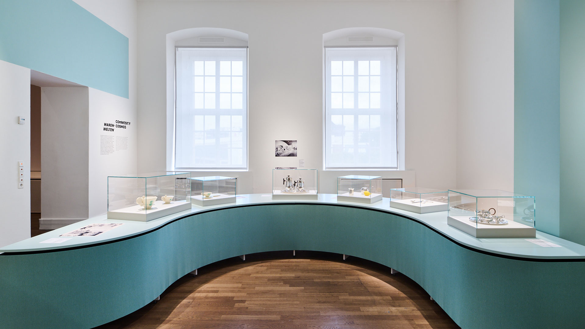 Raumansicht Ausstellung Widerstände. Vitrinen auf bumerangförmigem Podest. Hellblaue Atmosphäre.