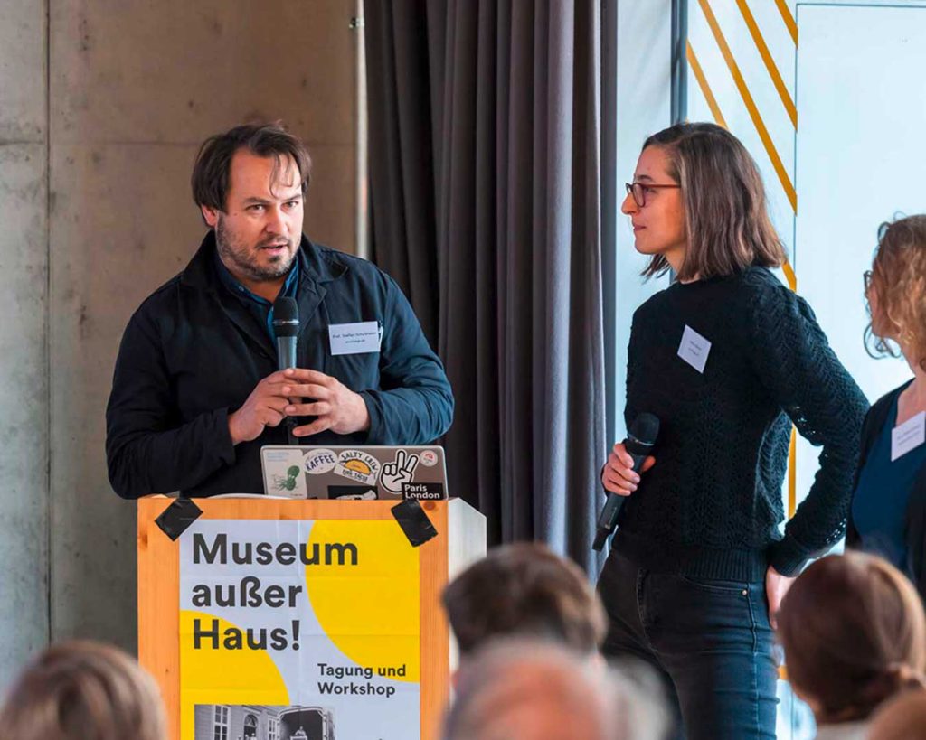 Zwei Personen moderieren auf der Bühne. Davor ein Plakat „Museum außer Haus!“