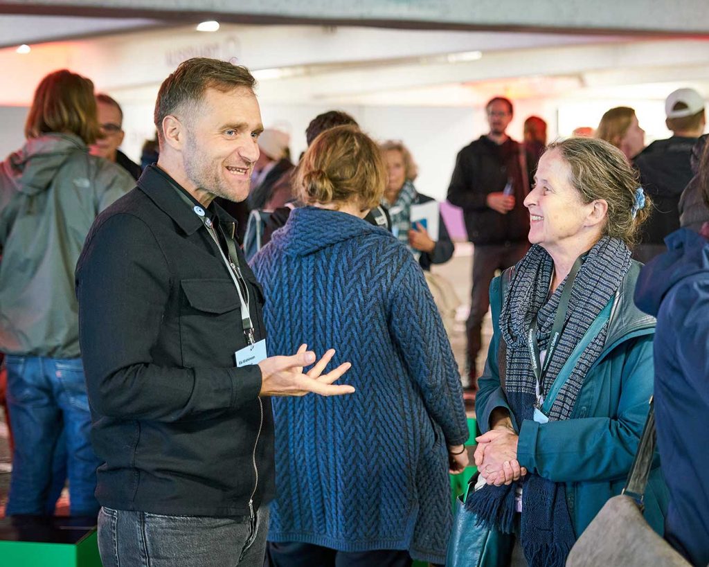 Zwei Personen im Dialog. Links gestikuliert eine Person, rechts hört eine aufmerksam zu und lächelt.