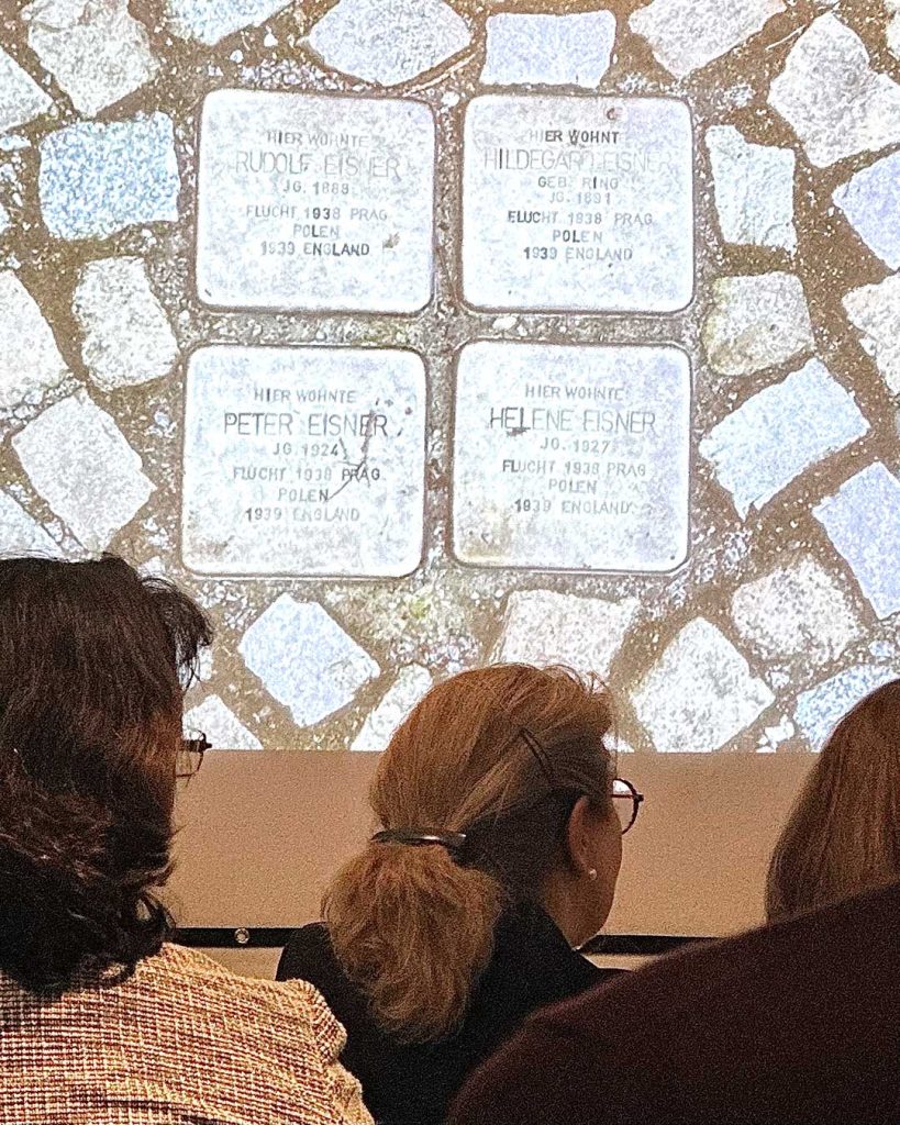Blick von hinten auf Publikum. Im Hintergrund eine Projektion mit Berliner Stolpersteinen.