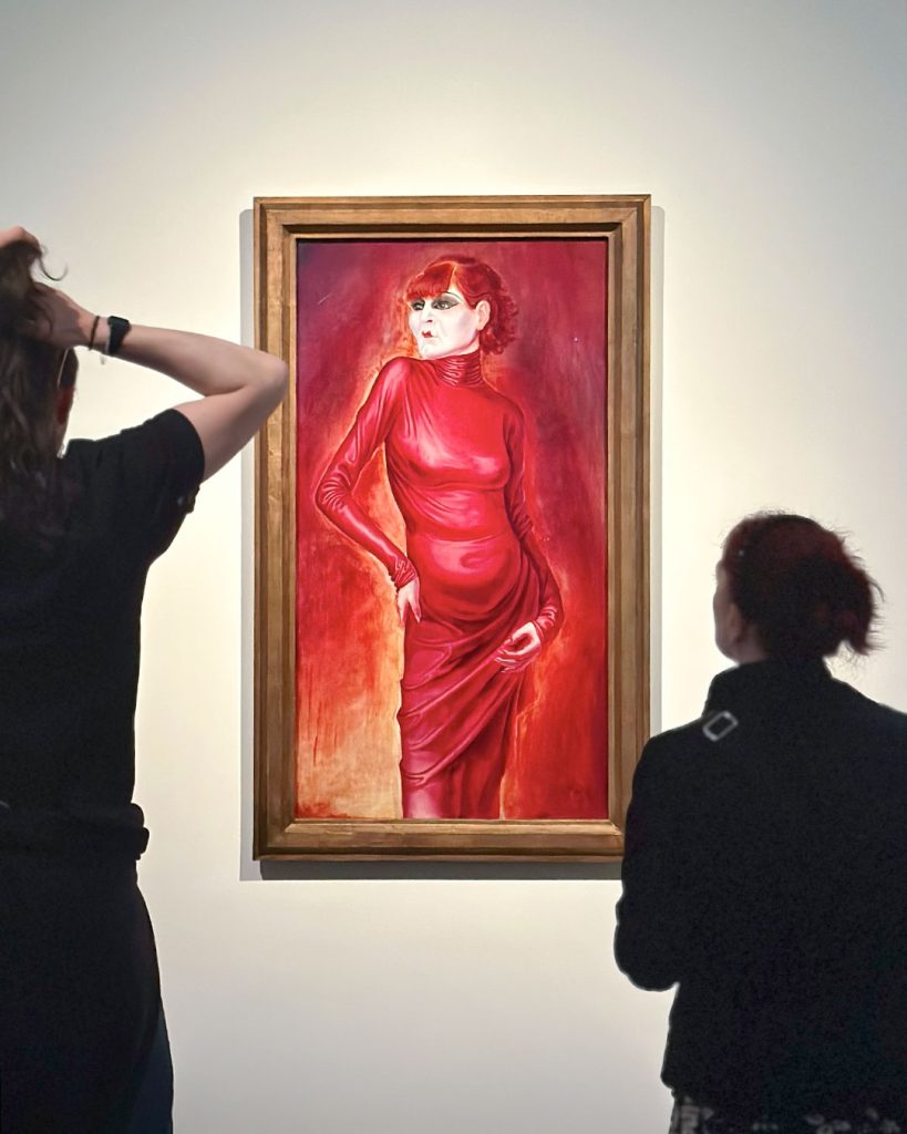 Blick zwischen zwei Personen hindurch auf Otto Dix’ Anita Berber. Rothaarige blass-weiße Person in hautengem roten Kleid vor rotem Hintergrund