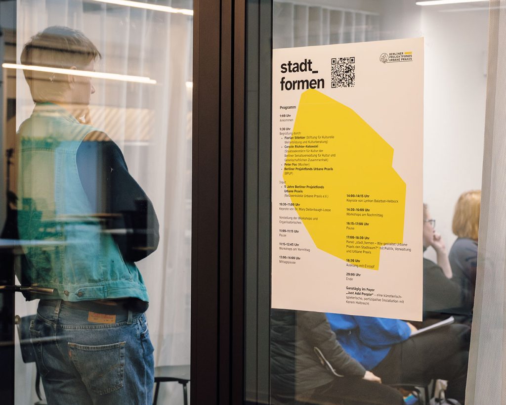 Konferenzprogramm Plakat auf Glasscheibe am Zugang zum Konferenzraum. Eine Person lehnt am linken Seitenrand und hört aufmerksam zu